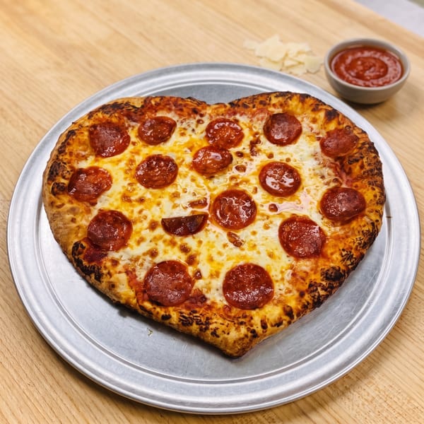 Heart-shaped pepperoni pizza on a metal tray with a side of marinara sauce and parmesan cheese.