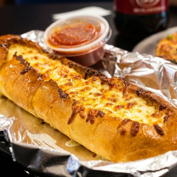 Cheesy garlic bread in foil with a side of marinara sauce, and a pizza in the background.