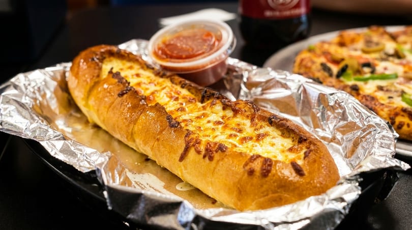 Cheesy garlic bread in foil with a side of marinara sauce, and a pizza in the background.