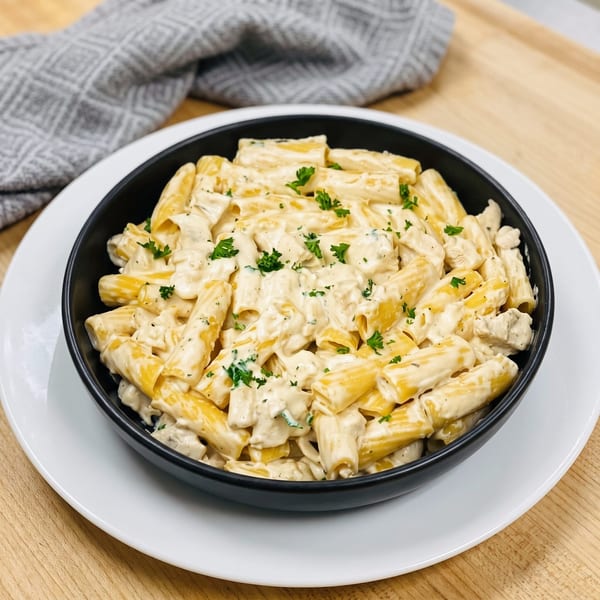 Creamy chicken rigatoni pasta with parsley in a black bowl on a white plate and wooden table.